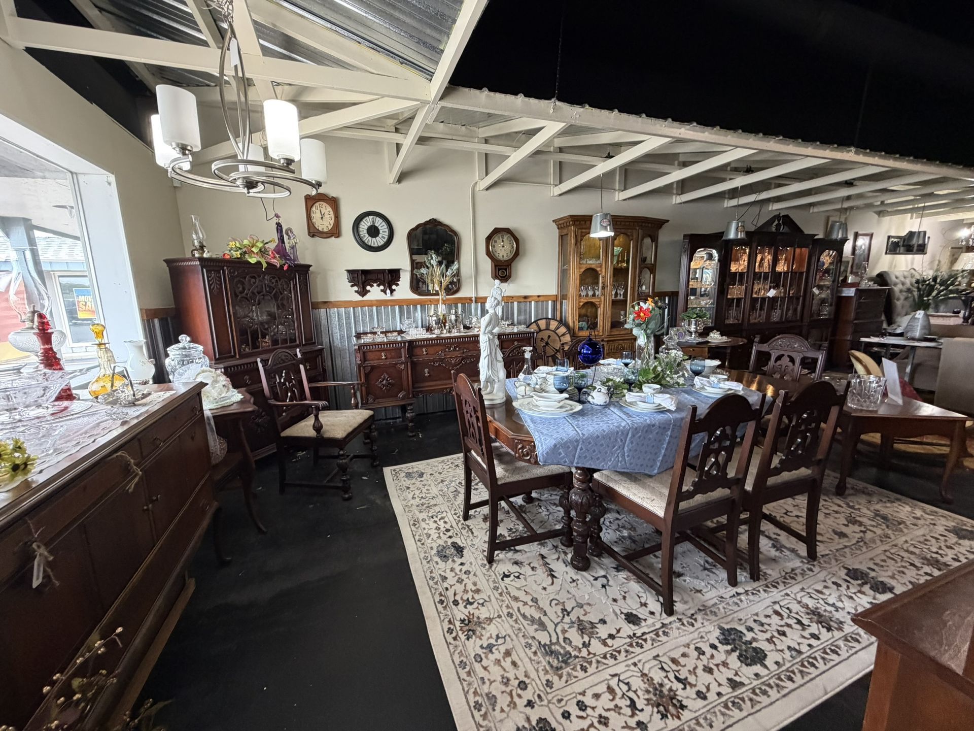 Antique-filled dining room with wooden tables, chairs, cabinets, and a patterned rug under exposed beams