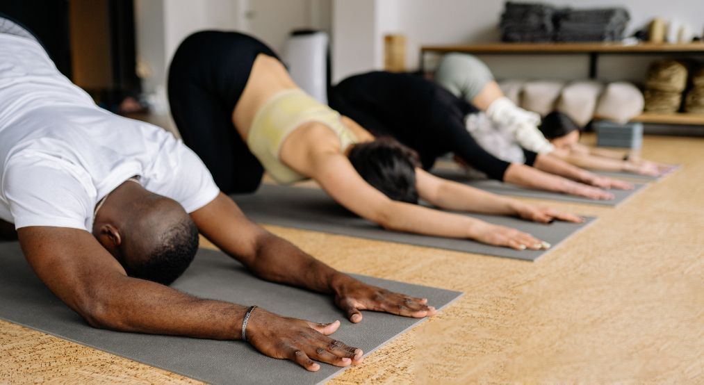 People in a yoga class stretching in a child's pose, on mats, with a neutral setting.