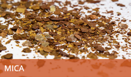 Brown mica flakes scattered on a white surface, with a label reading “MICA” at the bottom.