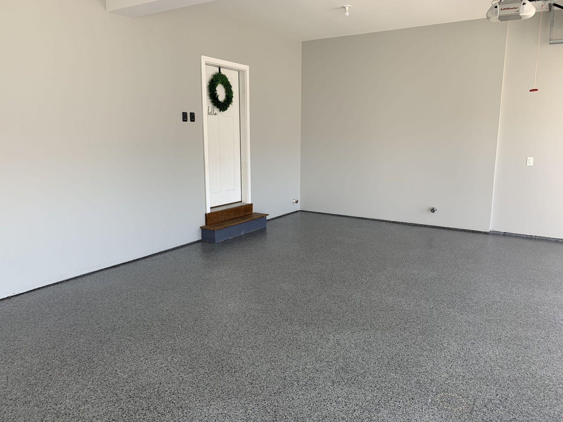 Empty garage with gray speckled floor and white walls, featuring a door with a wreath.