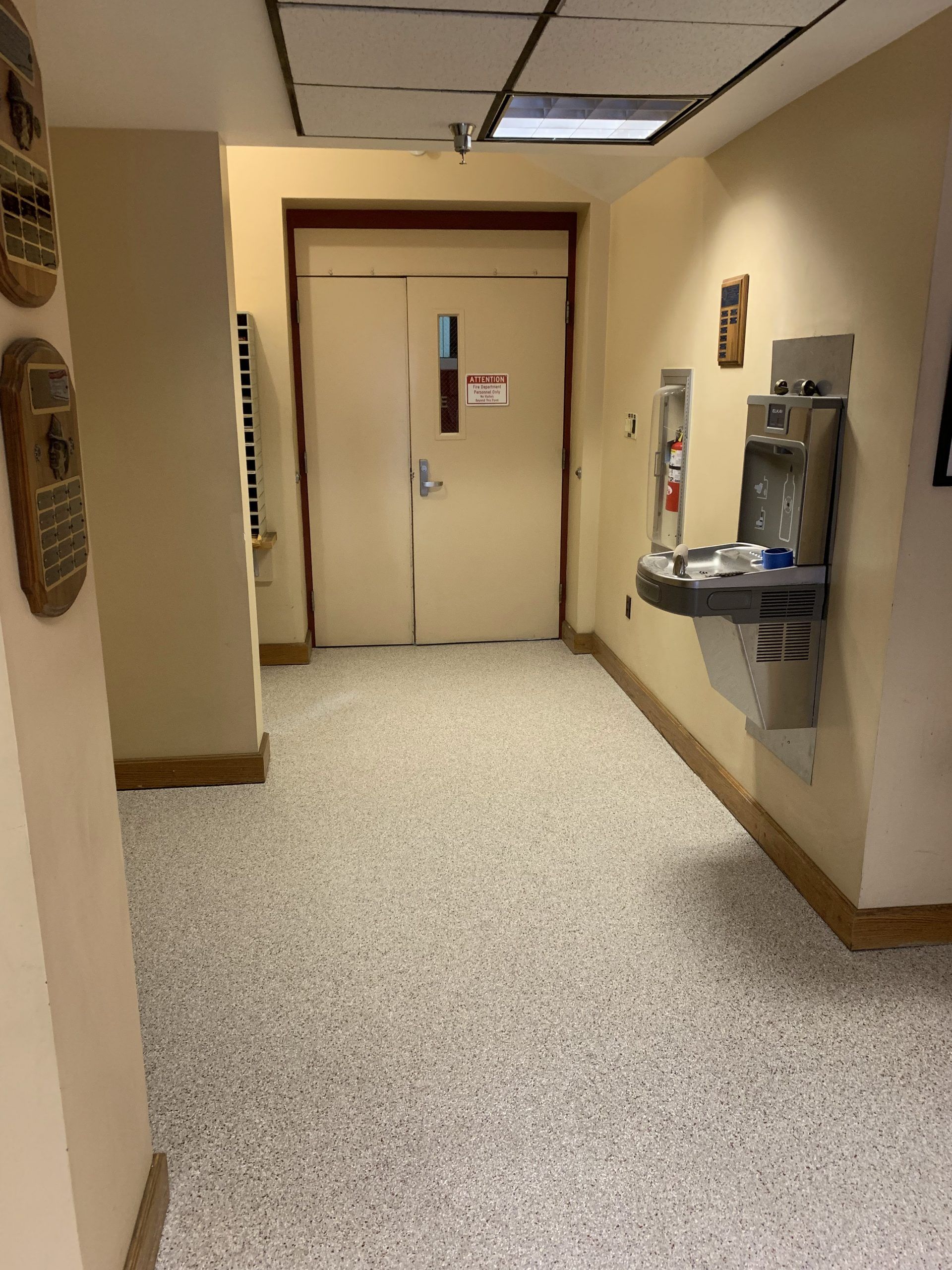 Office hallway with beige walls, speckled floor, closed double doors, and a water fountain on the right.
