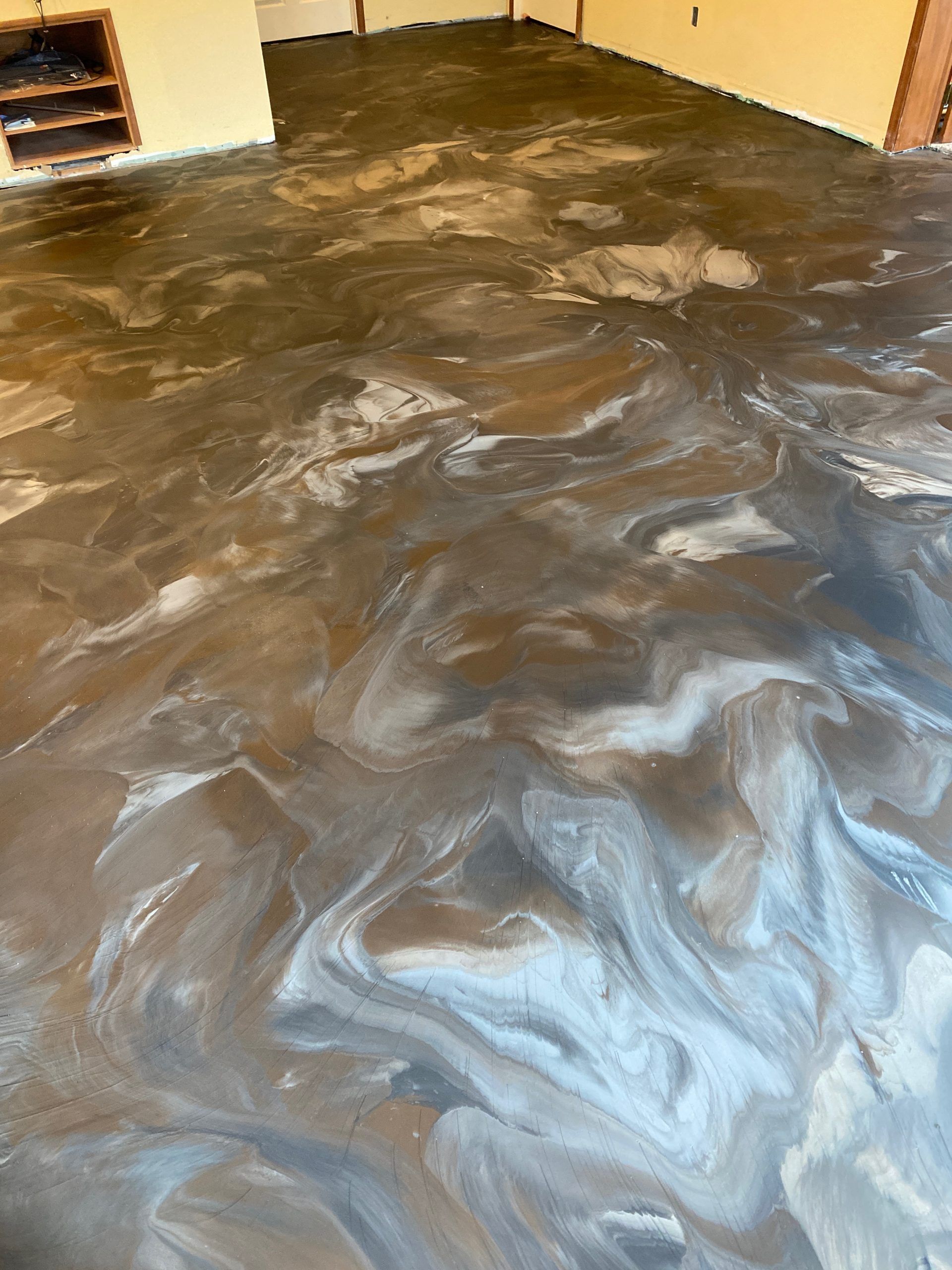 Swirled dark epoxy floor finish in an unfinished room with wood trim and a brick wall.
