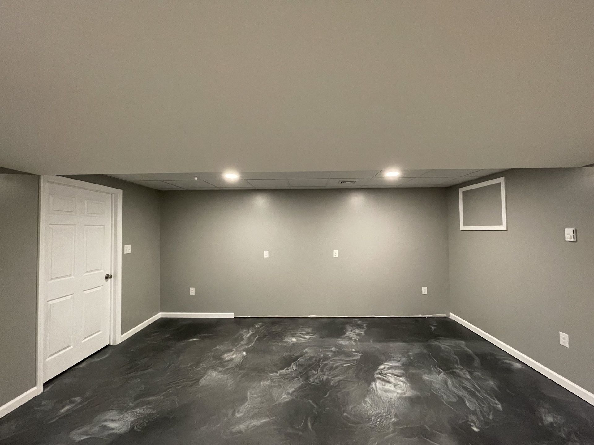 Empty gray finished basement room with dark floor, white door, recessed lights, and small window opening