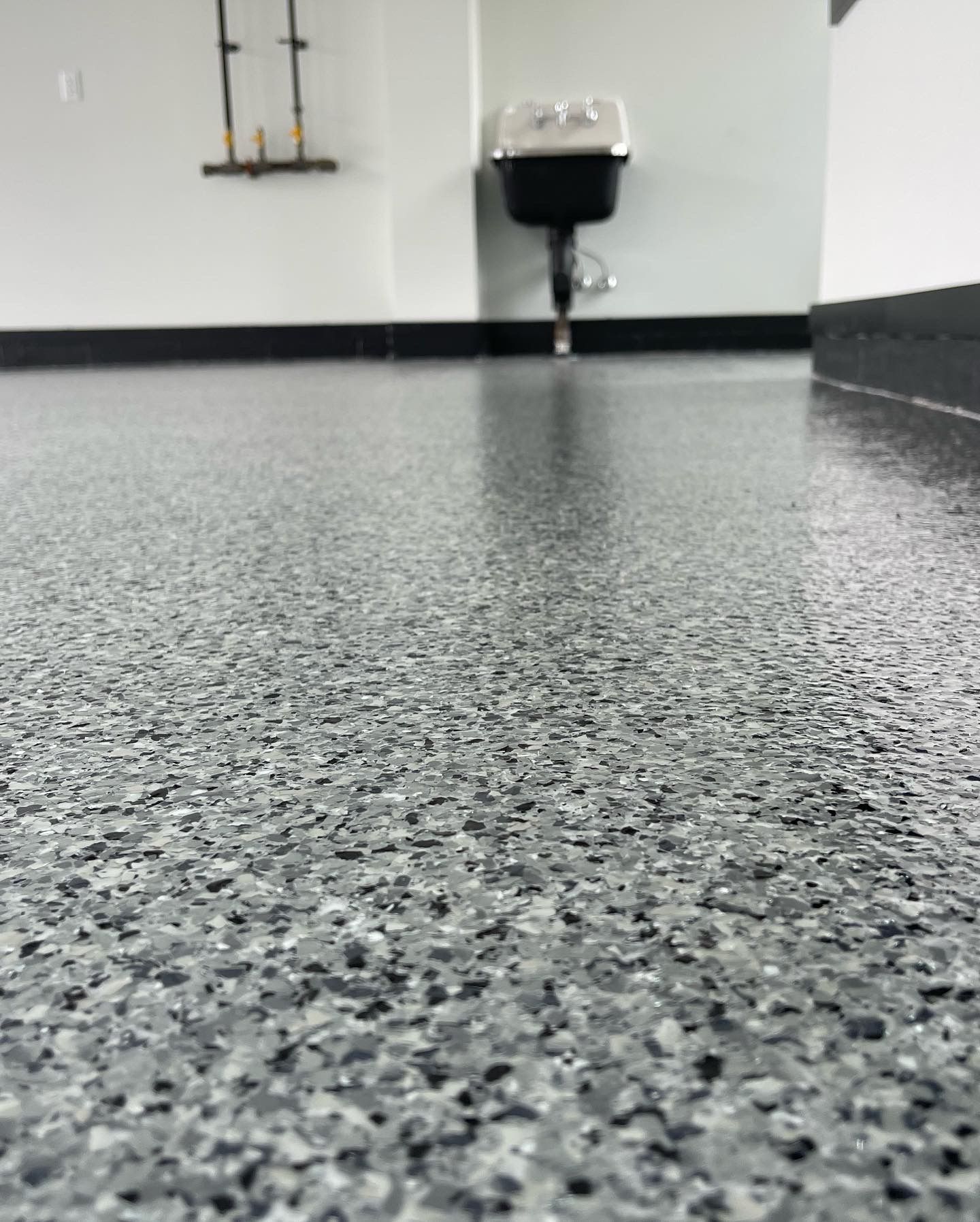 Low-angle view of a speckled gray floor with a wall sink and pipes in the background.