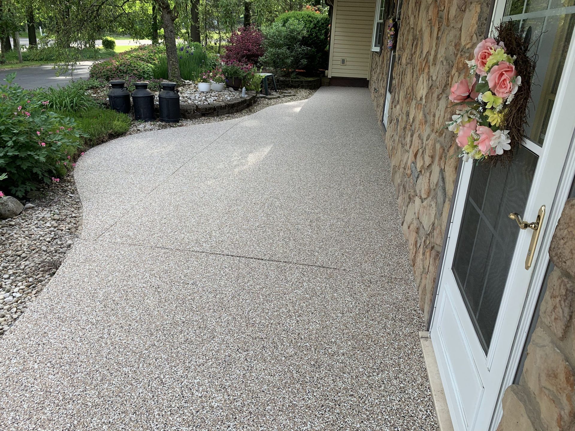 Gravel walkway beside a house with stone siding, a white door, and flower wreaths, bordered by garden beds.