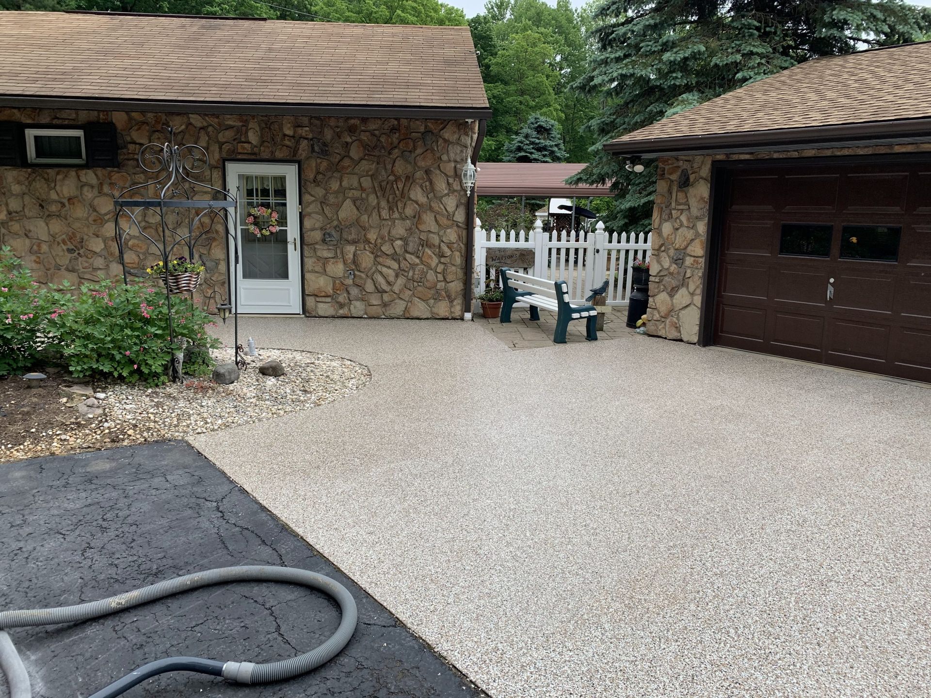 Driveway between two stone buildings with a garage, white fence, and garden hose on the ground.