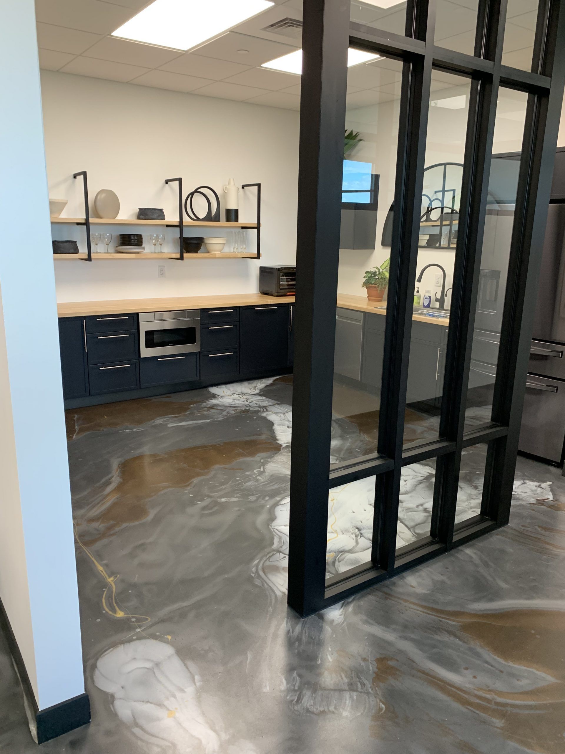 Modern office kitchen with black cabinets, open shelving, and glass partition; polished concrete floor.