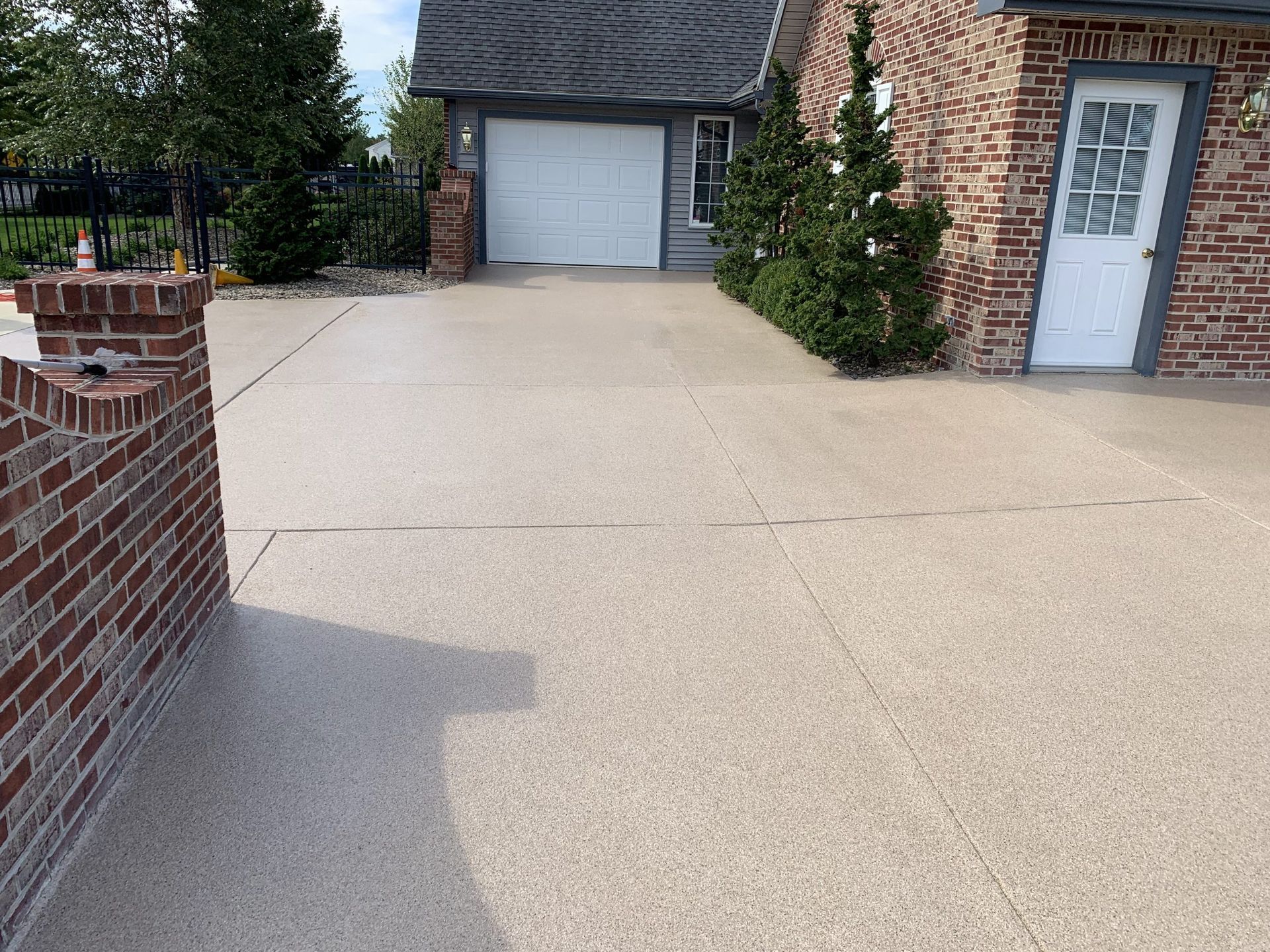 Brick house driveway with a large concrete patio and garage doors, bordered by small shrubs and a side door