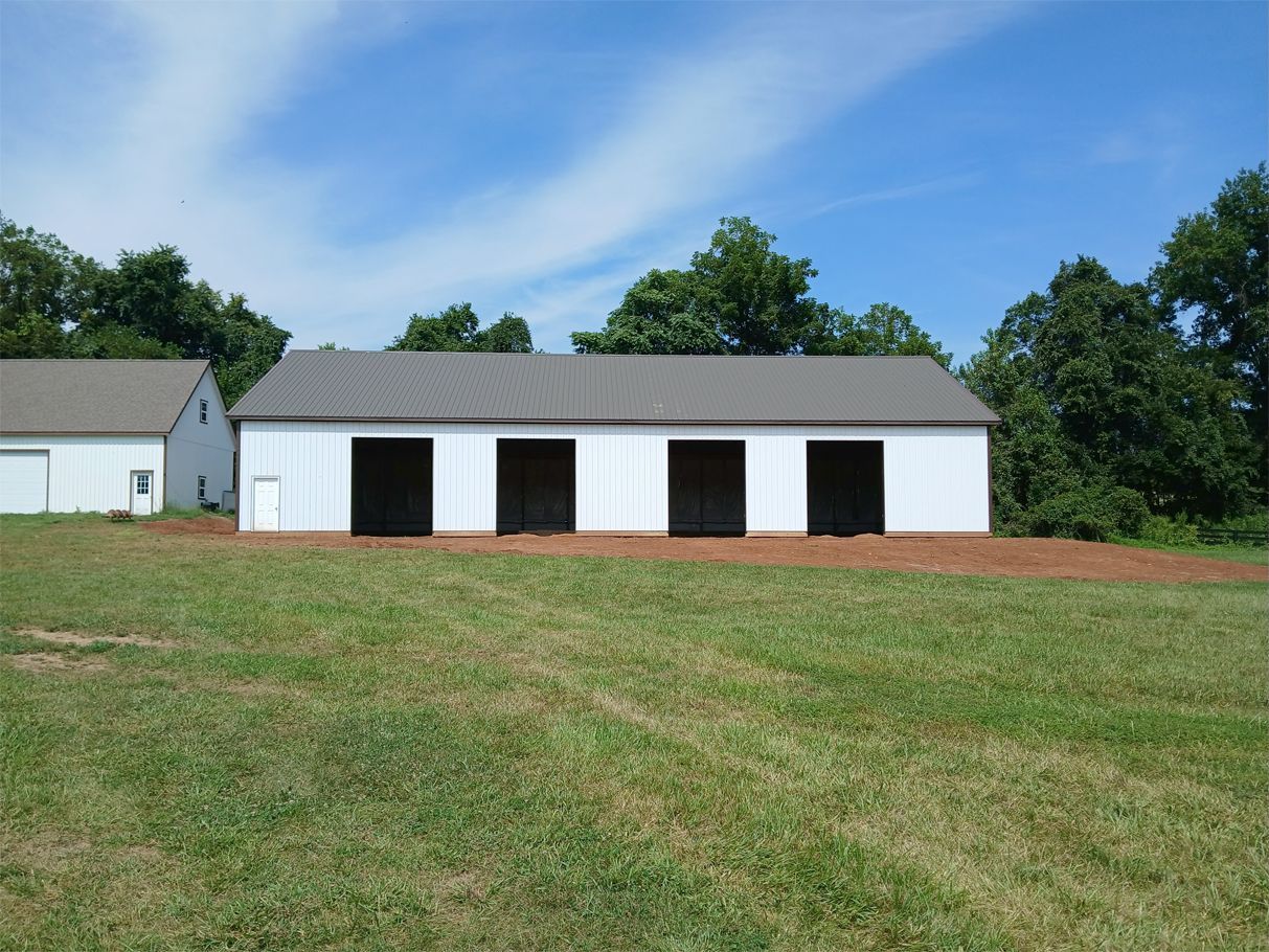 White barn with black stalls, brown roof, green grass, and blue sky.