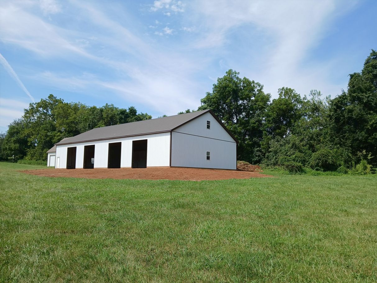 White barn with open stalls, brown roof, and green field under a blue sky.