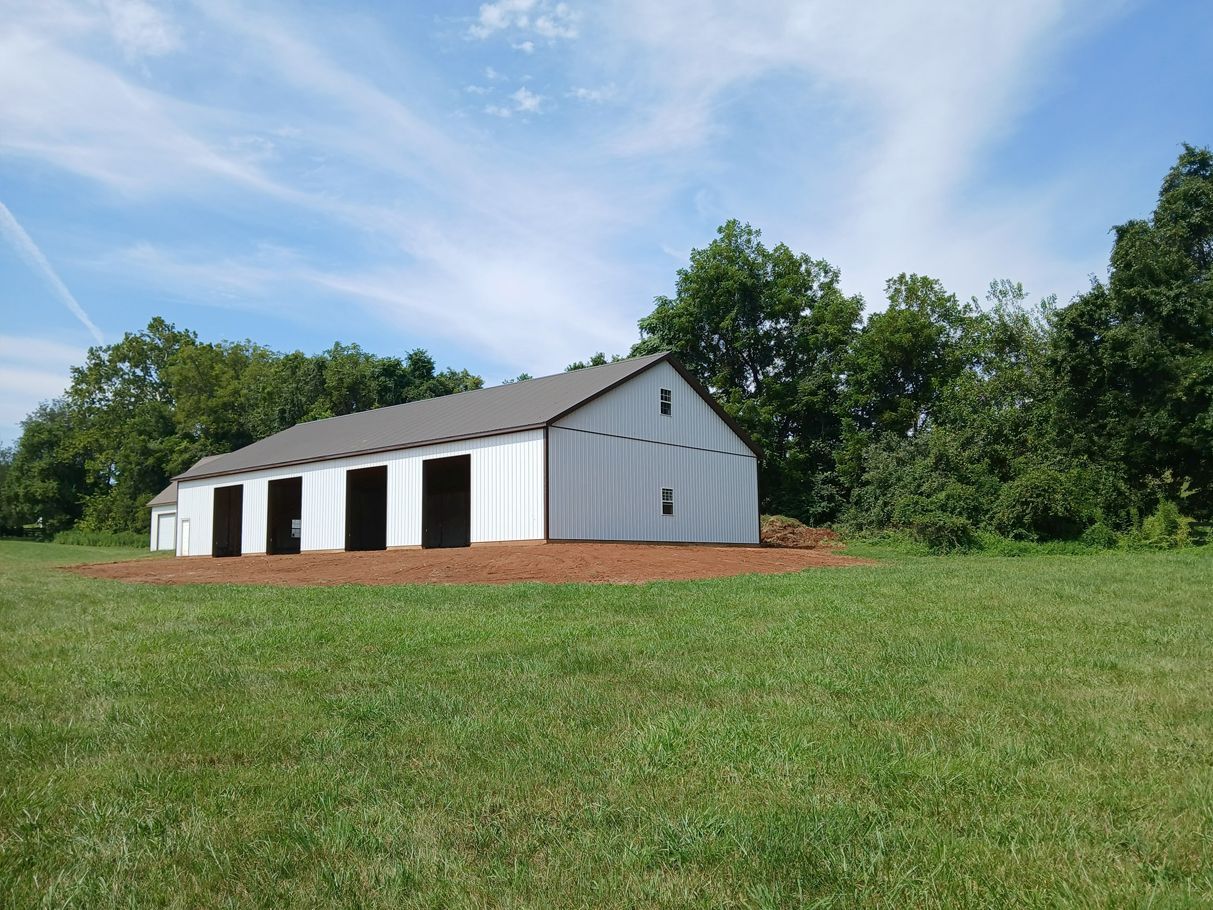 White barn with open stalls in a grassy field, trees in background under a blue sky.