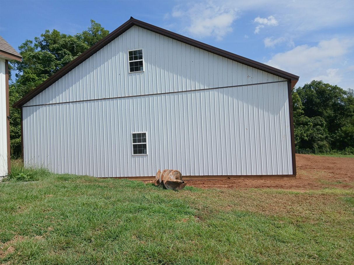 White barn with brown trim and wavy shadow on the side, set in a grassy field under a blue sky.