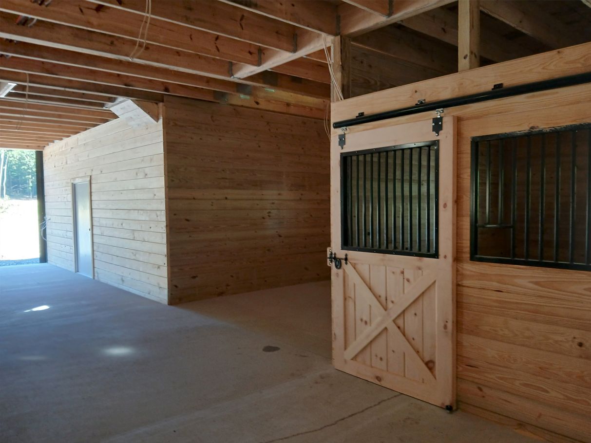 Interior view of a wooden barn with open stall door, showing concrete floor and unfinished ceiling.