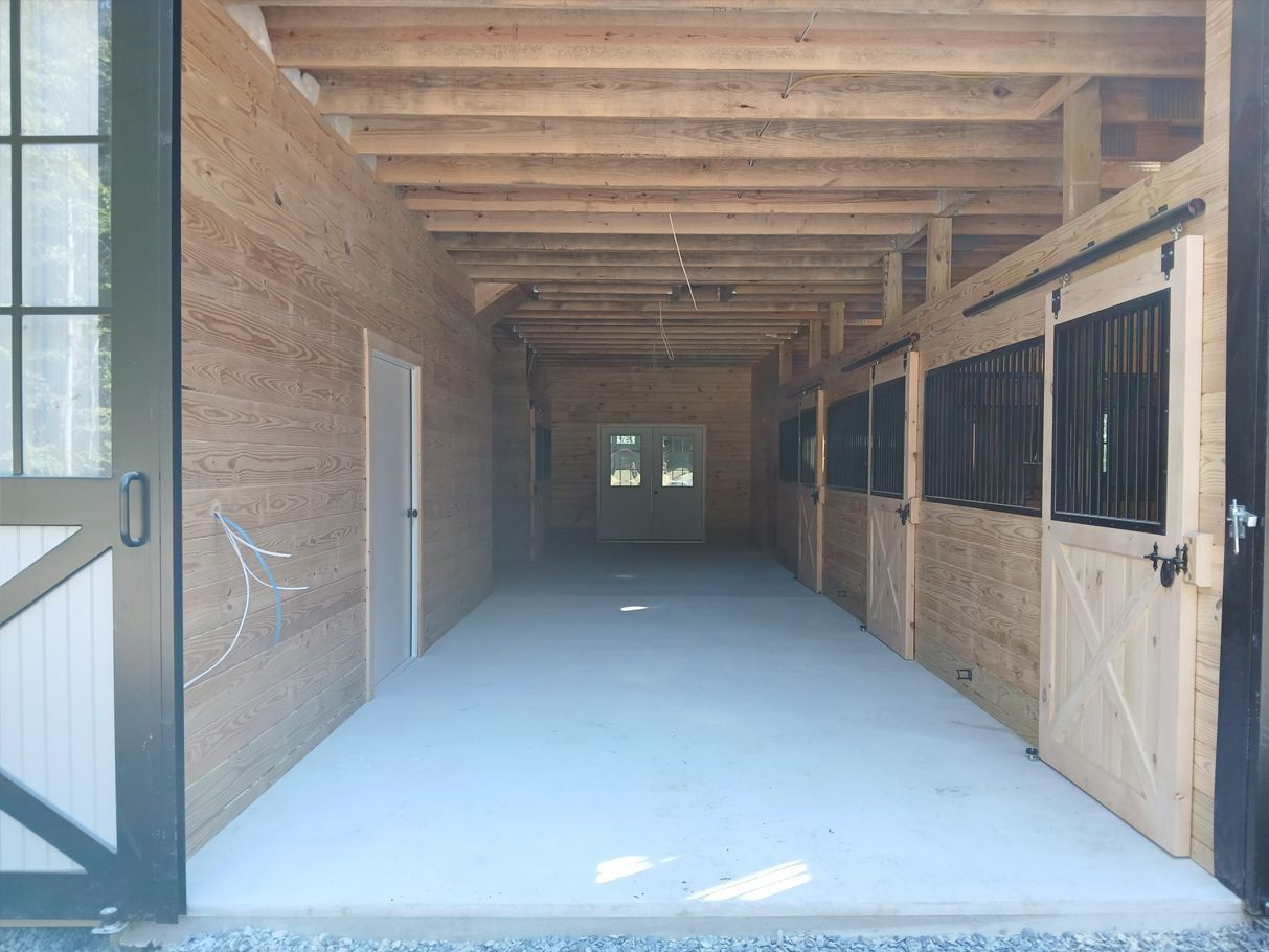 Interior of a horse stable with light-colored stalls, concrete floor, and open doorway.