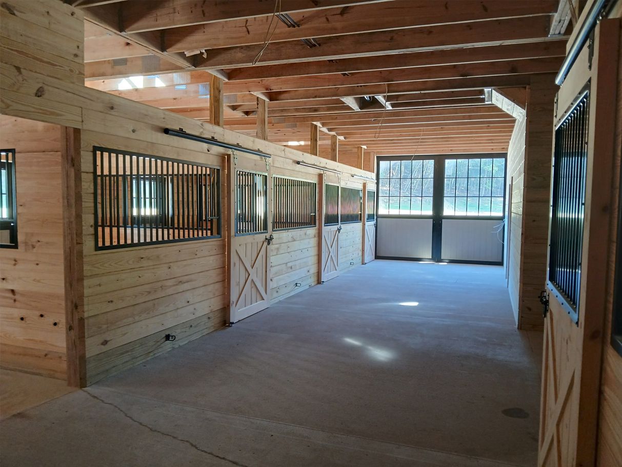 Interior of a wooden barn with horse stalls. The floor is concrete. Sunlight streams through a large window.