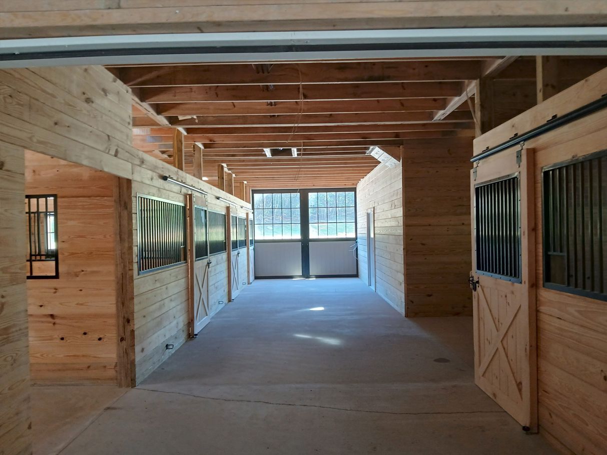 Interior of a horse stable with wooden stalls and sliding doors. Sunlight streams through the windows at the end of the aisle.