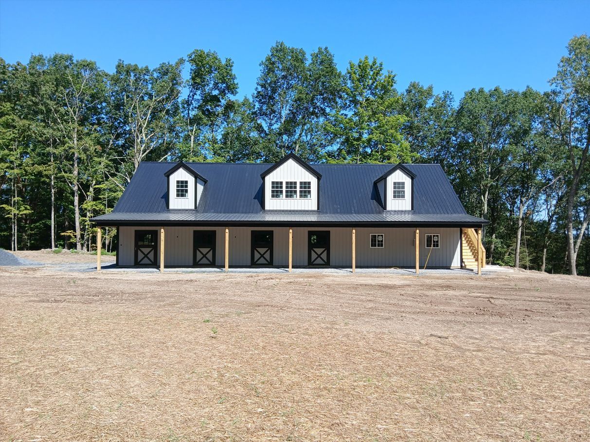 Horse barn with a black roof, gray siding, and a row of horse stalls under a covered porch, against a backdrop of trees.