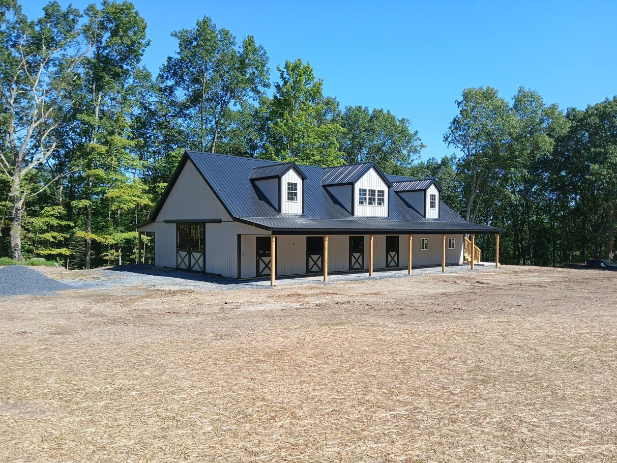 White barn with black roof, dormers, and horse stalls, set against a backdrop of trees and a clear blue sky.