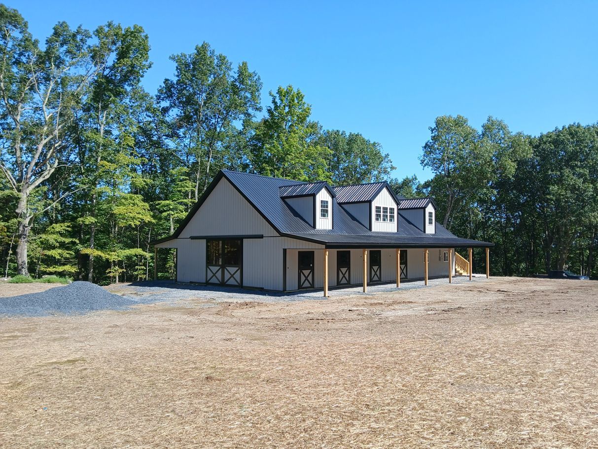 White horse stable with black roof and dormers; set in a clearing on a sunny day.
