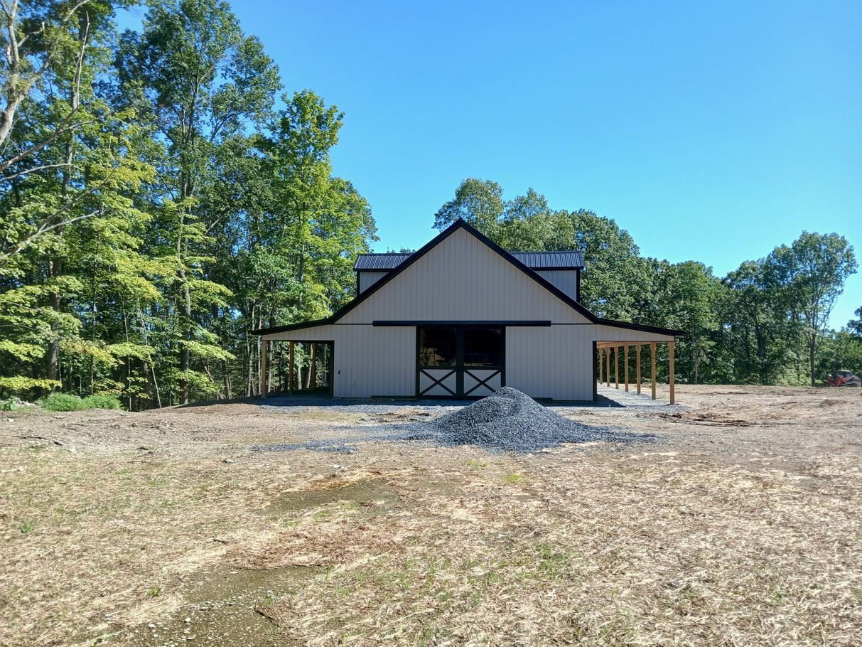 Tan barn with black roof and doors, surrounded by trees and a blue sky.