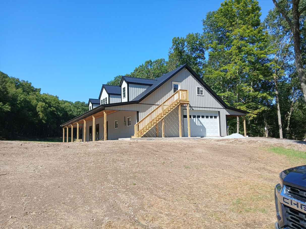 Two-story barn-style building with porch, garage, and exterior staircase, set against a blue sky and treeline.