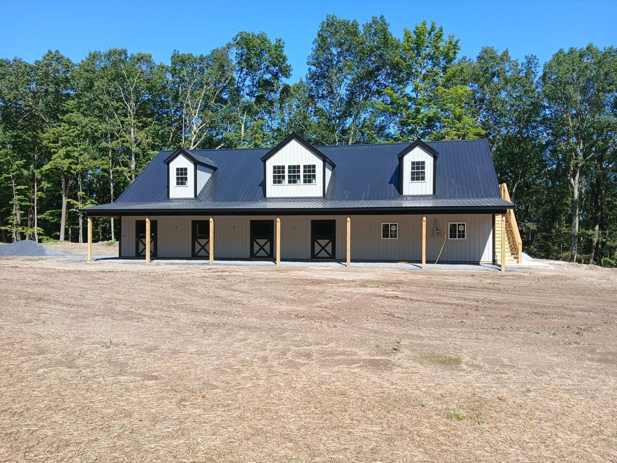 Horse barn with black roof, white dormers, tan walls, and black doors; set in field against a treeline.