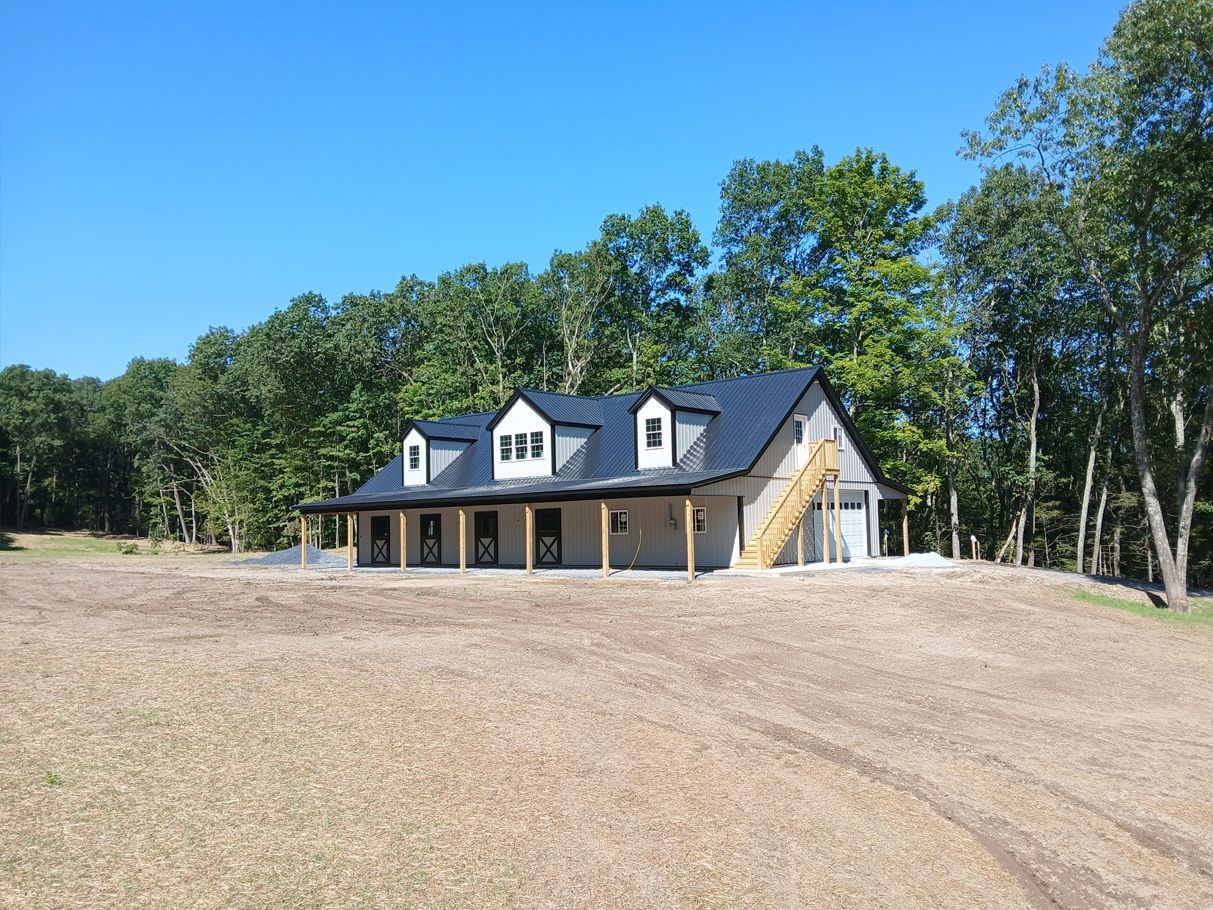 Gray and white barn-style building with dormers, black roof, and a garage. Set in a clearing near trees.