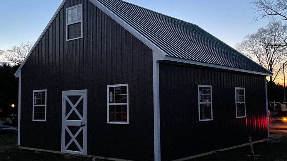 A black barn with white trim and windows is sitting in the middle of a field.