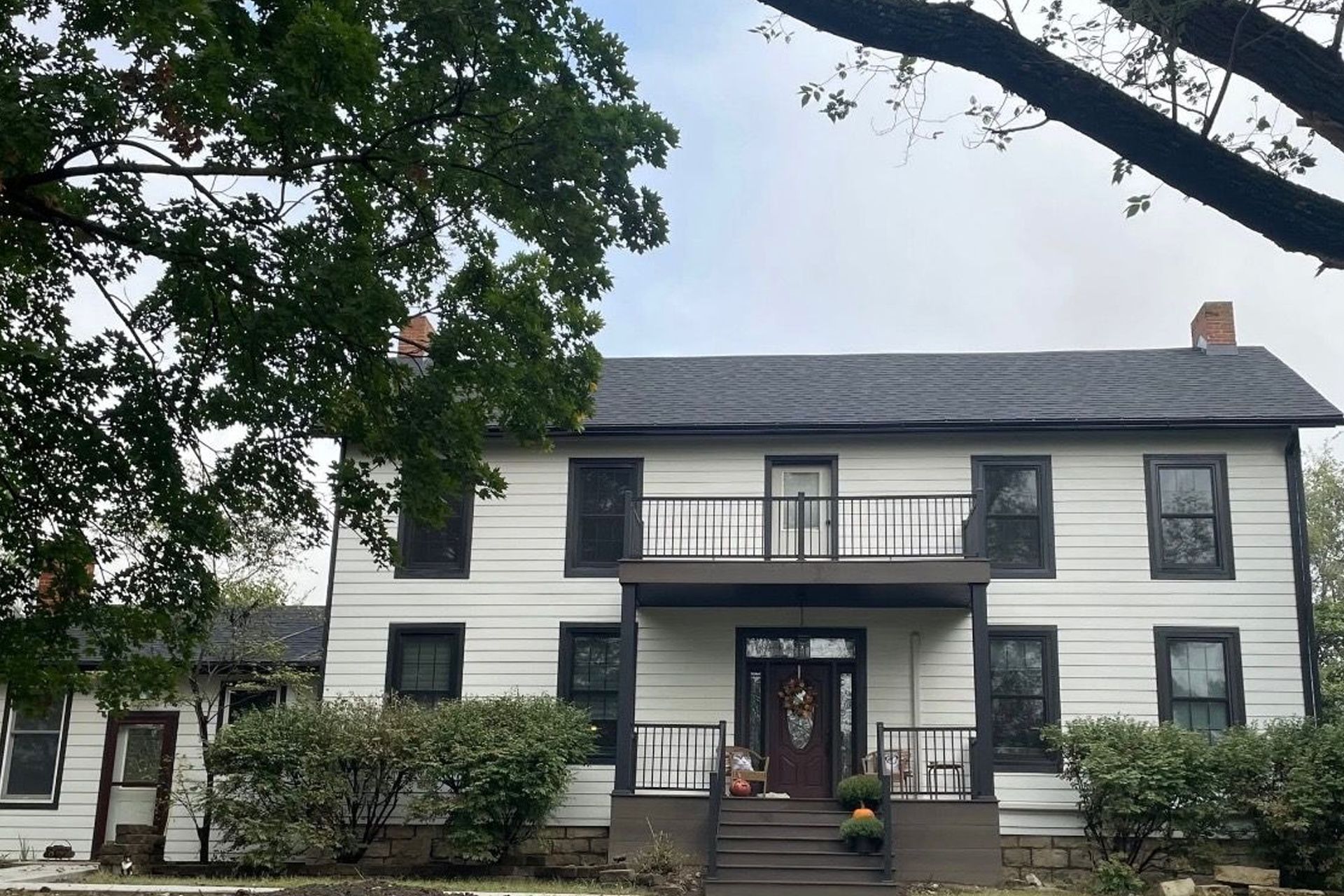 Two-story white house with black trim, balcony, and dark roof under a cloudy sky; tree in the foreground.