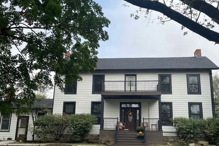 Two-story white house with black trim, balcony, and dark roof under a cloudy sky; tree in the foreground.
