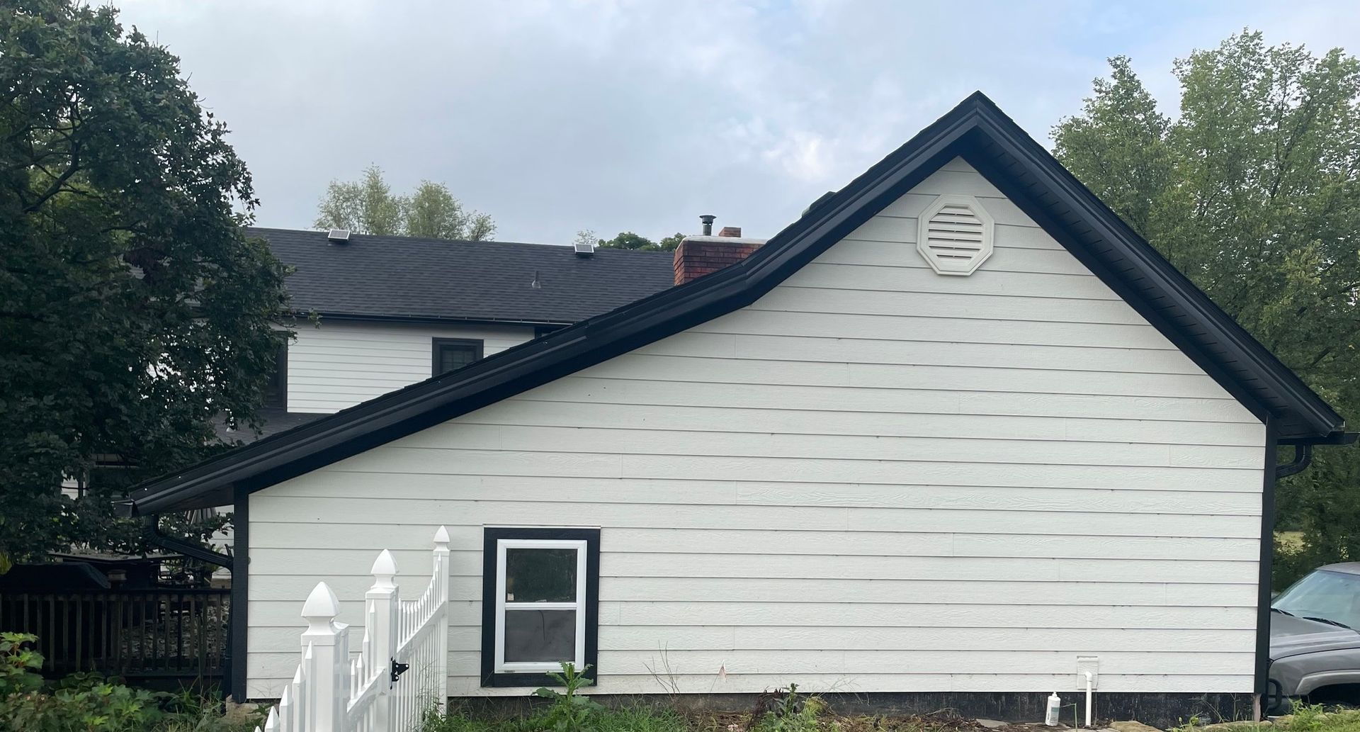 White house with black trim and roof, small window, a fence in front. Cloudy sky.