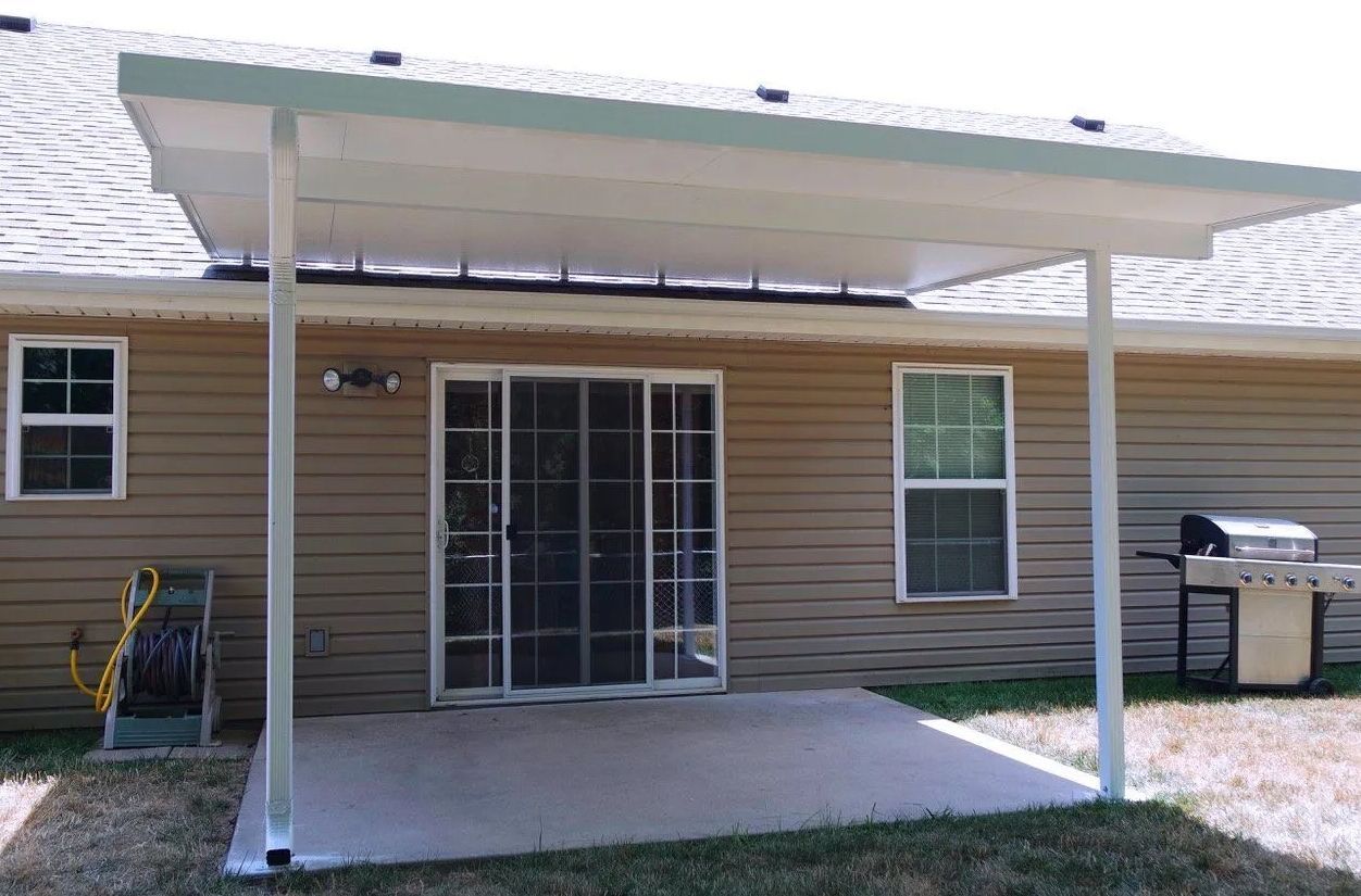A white patio cover extends over a concrete patio next to a beige house with a sliding glass door.