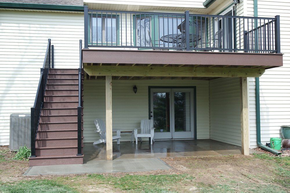 Two-story deck with black railing and stairs, brown decking, white house, patio with chairs.