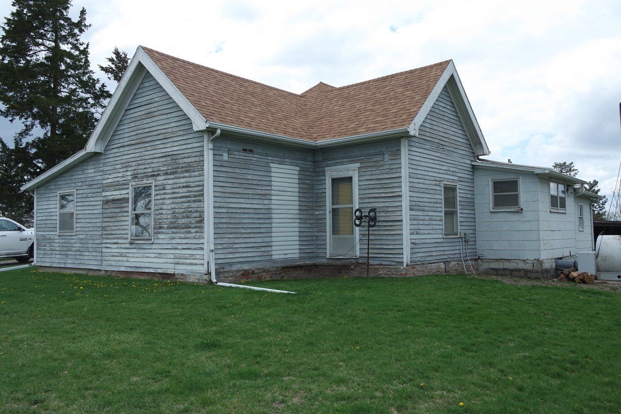 Weathered, two-story house with peeling paint; brown roof, green grass, and a car in the yard.