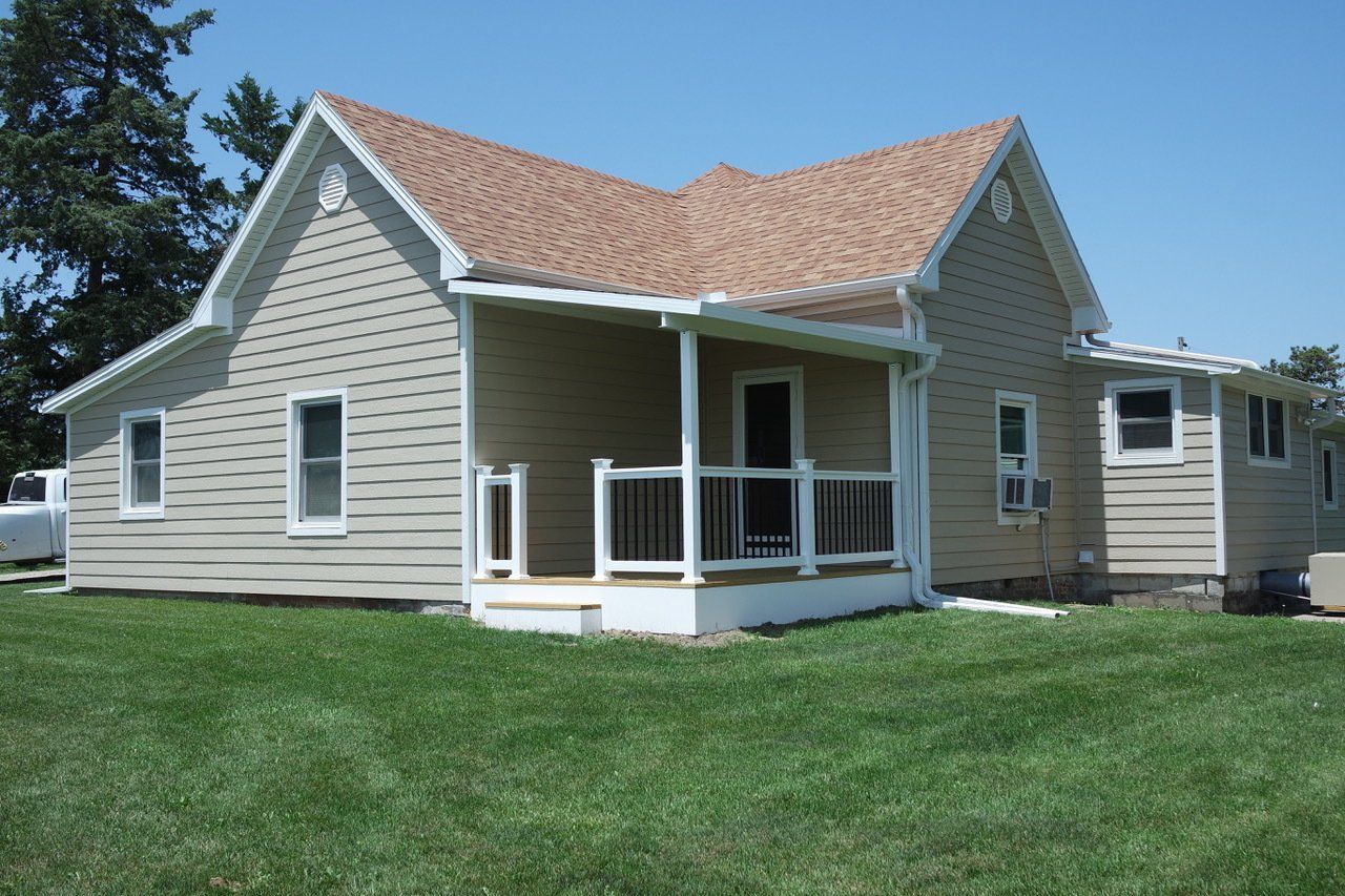 Beige house with brown roof and white trim, small porch, green lawn, and clear blue sky.