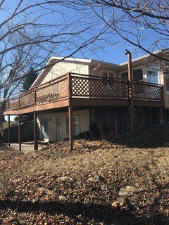 Wooden deck with lattice railing attached to a beige house on a slight slope. Brown dead leaves in foreground.