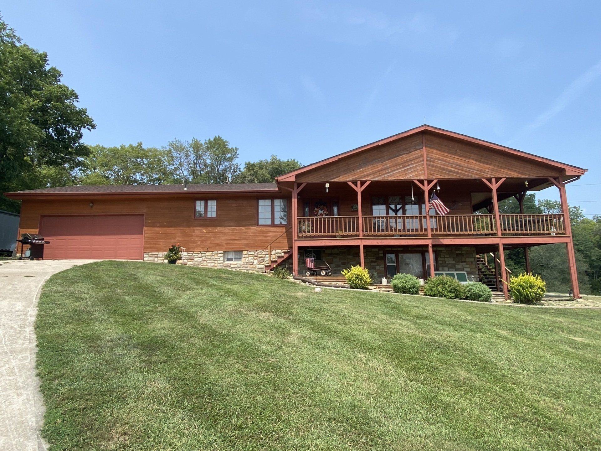 Brown house with a wraparound deck, stone and wood exterior, green lawn, and clear blue sky.