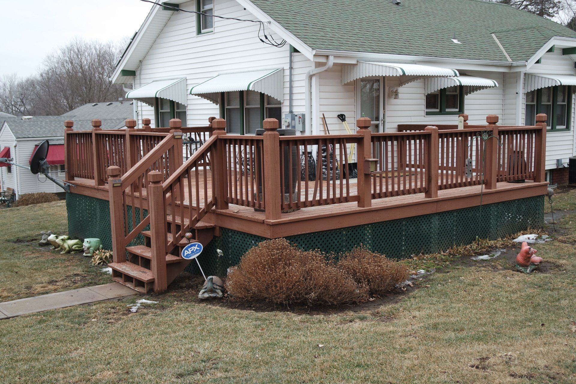 Wooden deck attached to a white house with brown trim.  Brown railing, stairs. Green lattice skirting. Cloudy day.