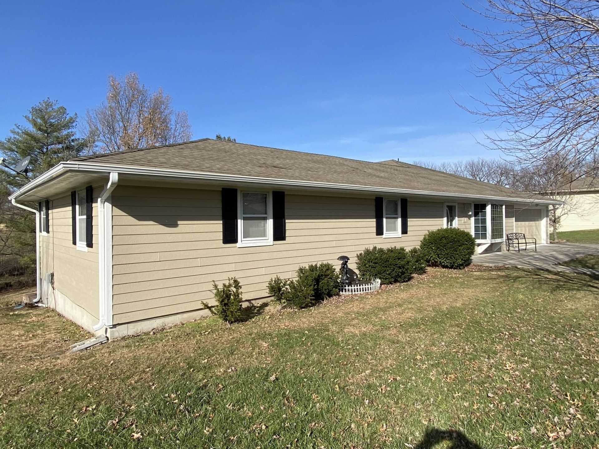 Beige ranch-style house with black shutters and a brown roof, on a grassy hill under a blue sky.