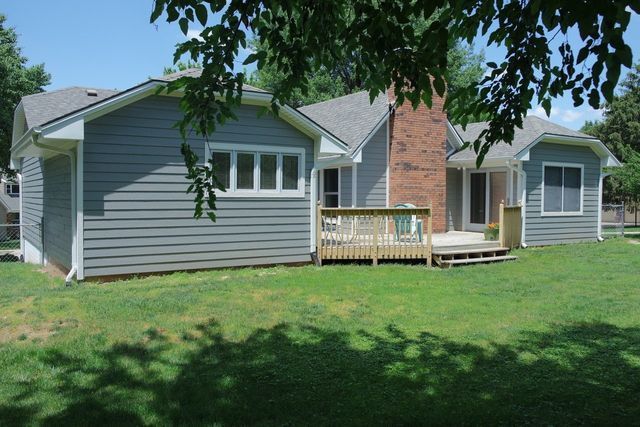 Backyard view of a gray house with a wooden deck, green grass, and tree shadows.