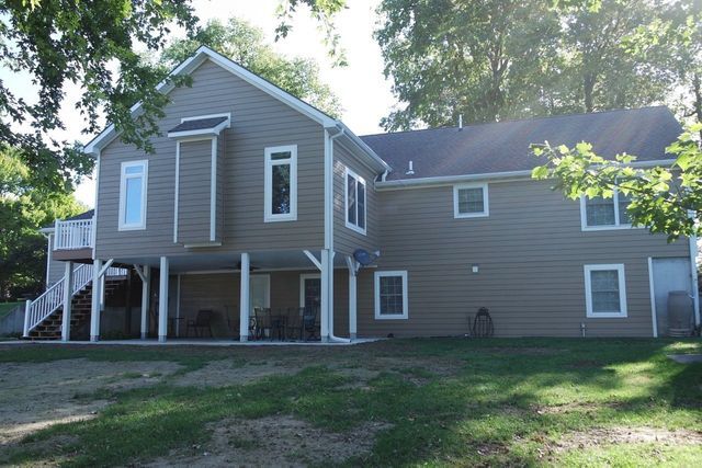 Two-story tan house with deck, windows, and white trim, seen from a grassy yard, trees overhead.