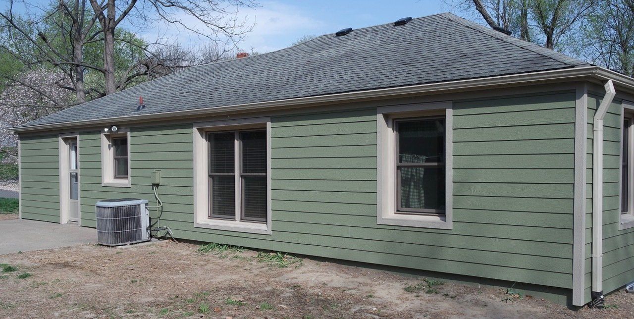 Green house with a grey roof, windows, and an AC unit outside.