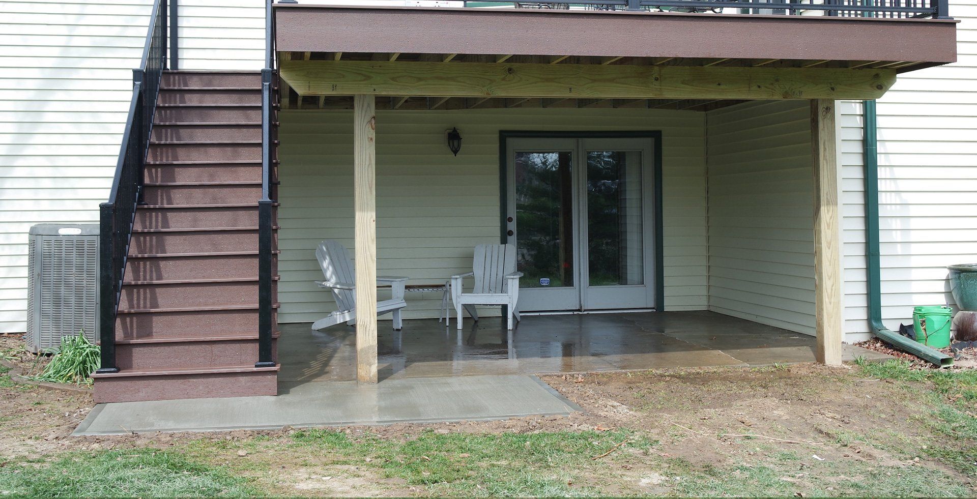 A deck with stairs leads to a covered patio with a door, chairs, and an air conditioner.