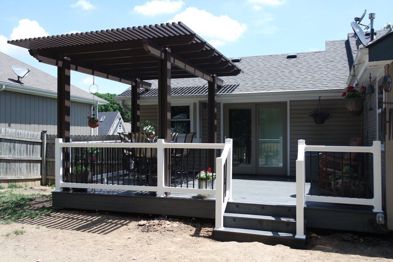 Backyard deck with pergola, white railing, and gray flooring. Brown wood pergola.