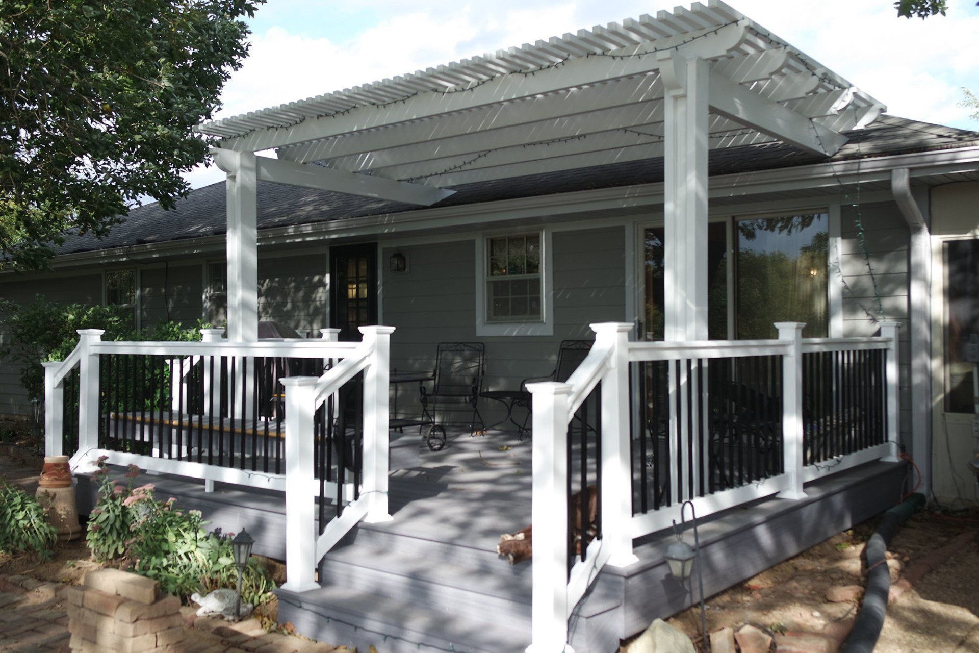 White pergola over gray deck with black railing, in front of a house.
