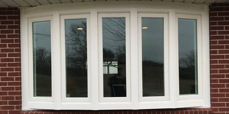 Bay window with white frames, set in a brick wall; glass reflects sky and trees.
