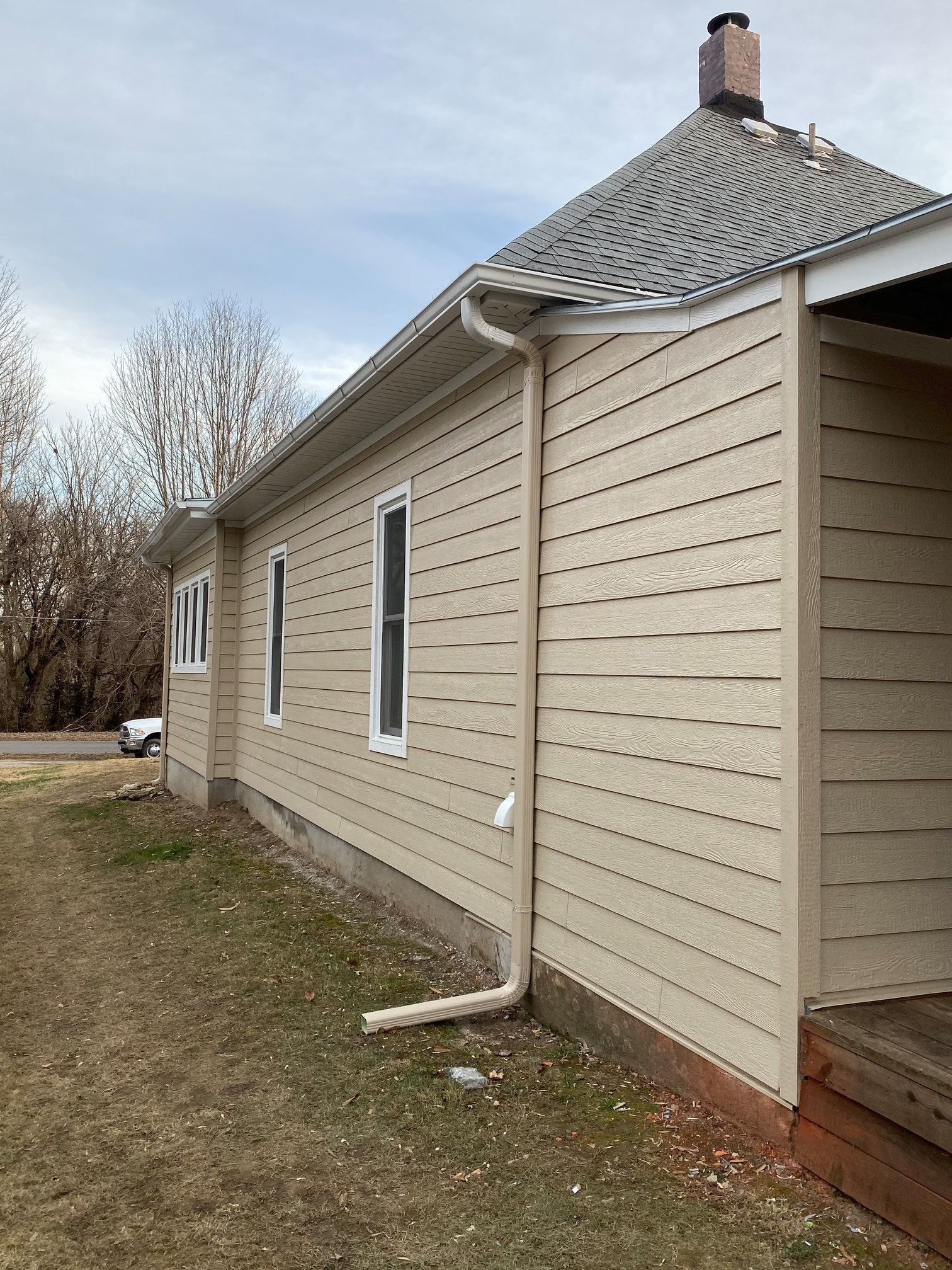 Tan-sided house with white window frames, a gutter, and brown roof. Outdoors, daytime, overcast sky.