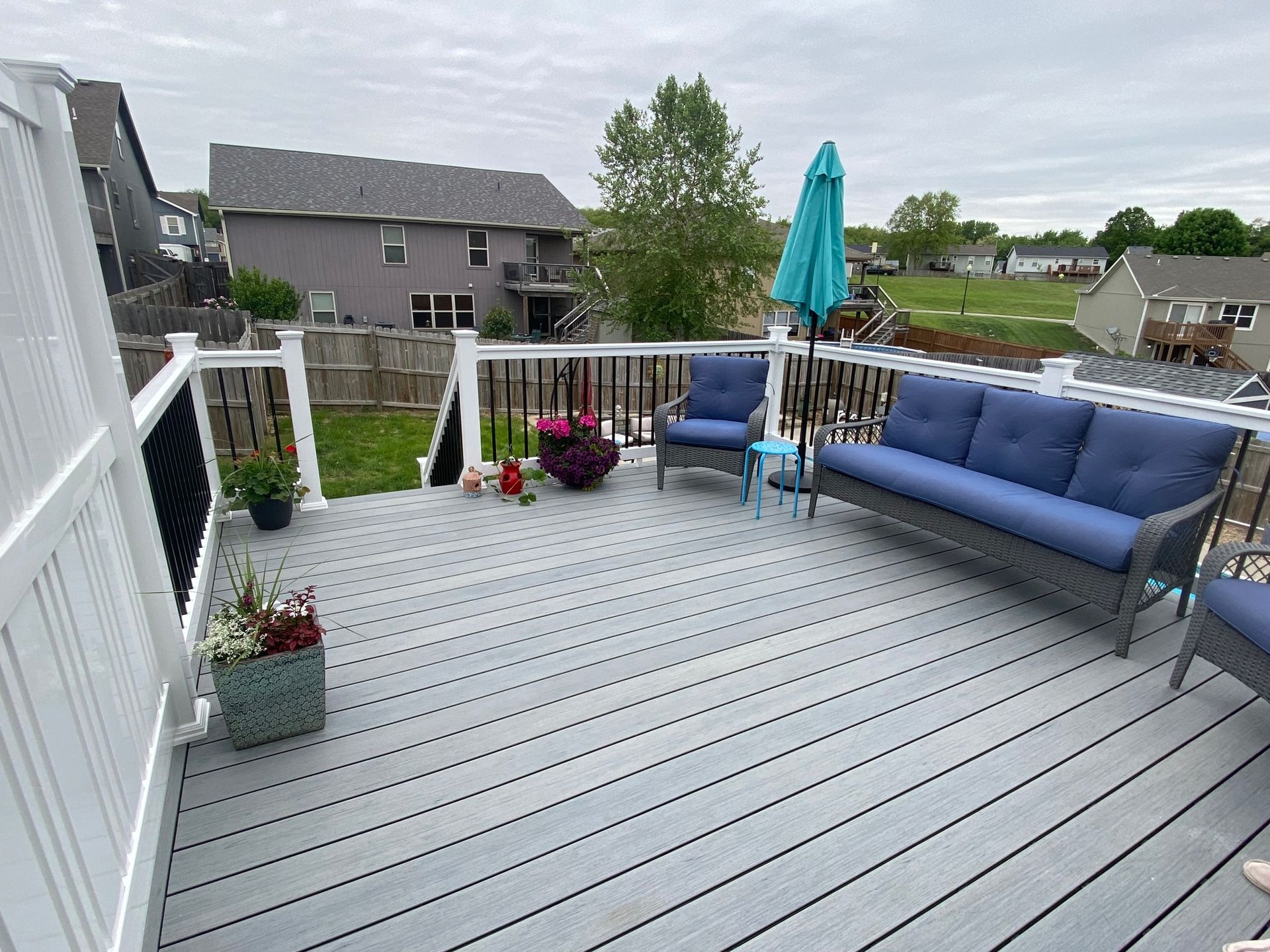 Grey composite deck with blue cushioned furniture and a teal umbrella in a backyard.