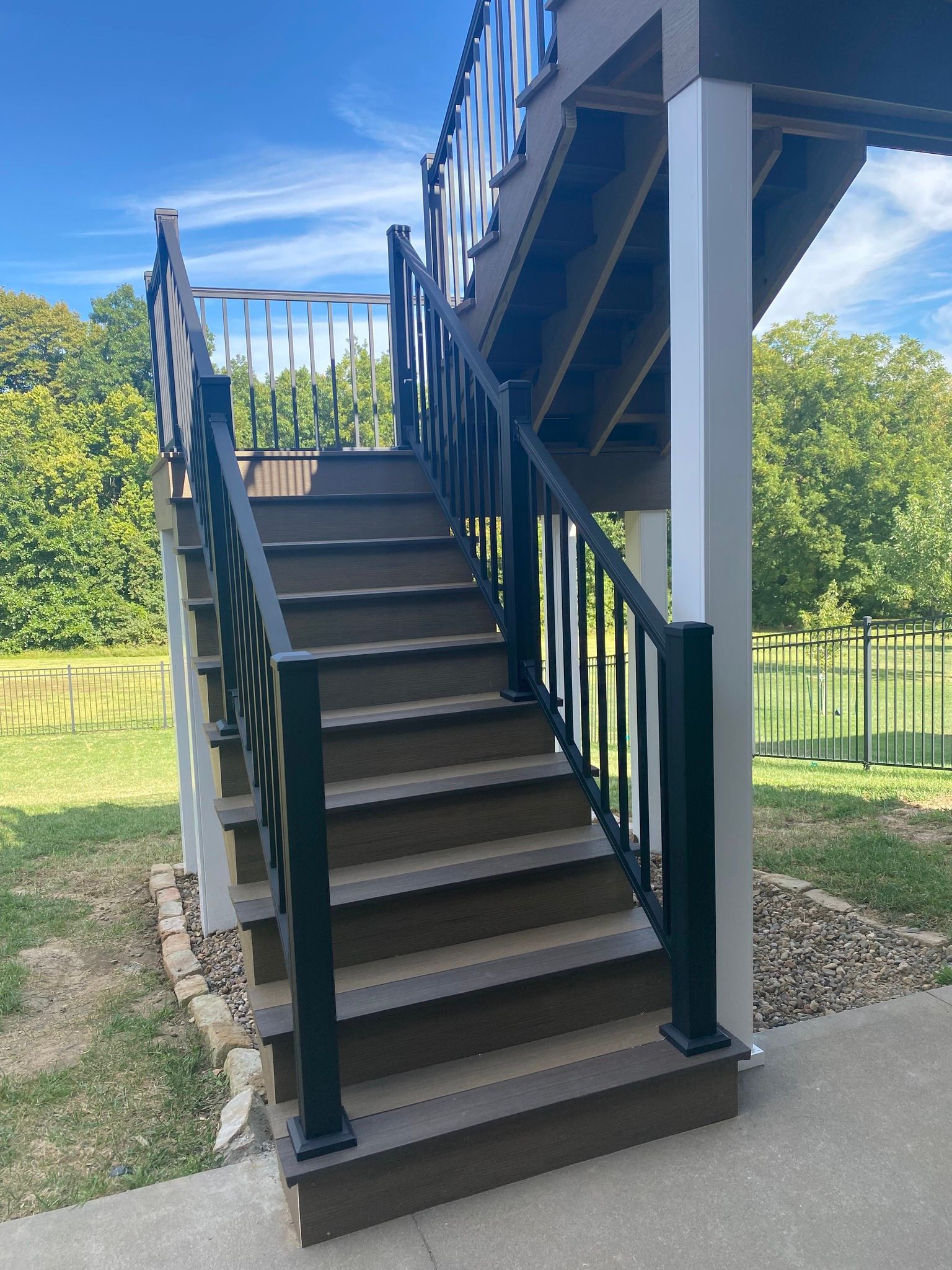 Stairs leading to a deck with black railings, against a backdrop of greenery and sky.