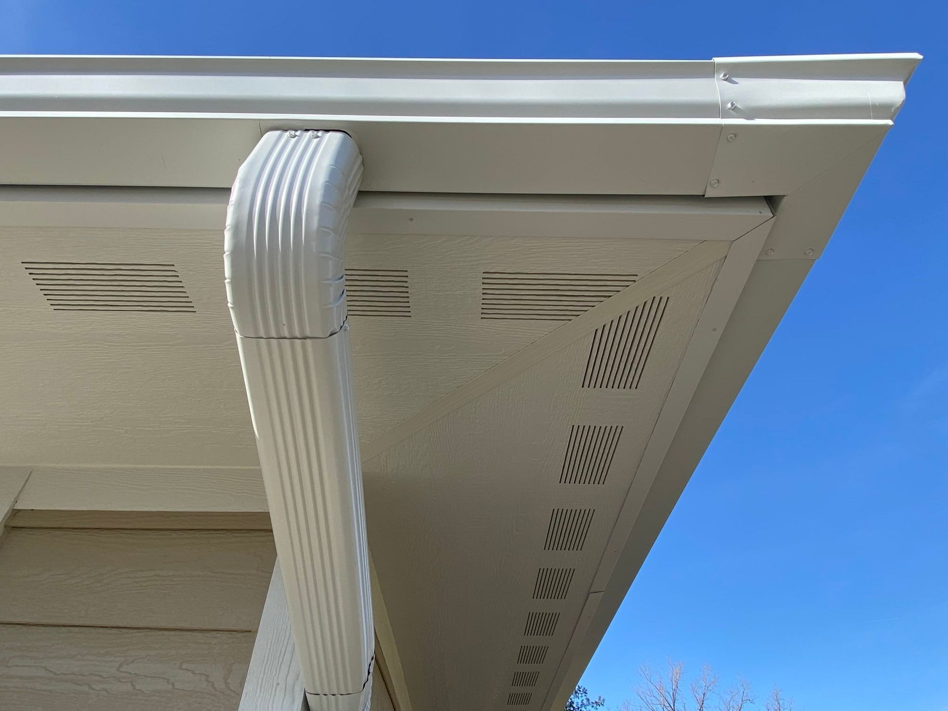 White gutter and downspout attached to a cream-colored soffit with ventilation slots, against a blue sky.